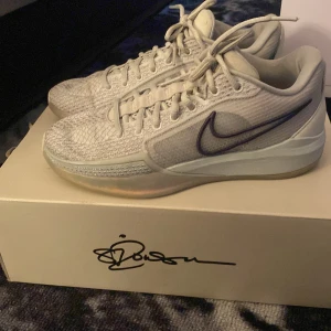 Nike Sabrina1  - Nike React sneakers i ljusgrått med mesh-överdel och snygga detaljer. Skorna har snörning, platt sula och ett stort svart Swoosh på sidan. Mönstrad yttersula och signatur på hälen. Perfekta för dig som gillar sportig och clean stil.