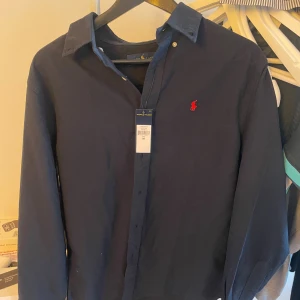 Mörkblå skjorta från Ralph Lauren - Snygg mörkblå skjorta från Ralph Lauren med klassisk krage och knappar framtill. Skjortan har lång ärm och den ikoniska röda Polo-loggan broderad på bröstet. Perfekt för dig som gillar stilrena och tidlösa plagg.