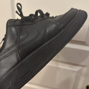 Nike Air Force 1 helsvarta sneakers - Säljer ett par klassiska Nike Air Force 1 sneakers i helsvart skinn. Skorna har rund tå, platt sula och snörning. Perfekta för dig som gillar en clean och tidlös look. Swoosh-loggan på sidan och diskreta detaljer gör dem till ett måste i garderoben.