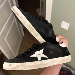 Golden Goose Superstar sneakers svart/vit - Golden Goose Superstar sneakers i svart skinn med vit stjärndetalj på sidan och vit häl. Klassisk låg modell med svarta skosnören och slitna, smutsiga vita sulor för den typiska vintage-looken. Snygg rund tå och platt sula. Kommer med originalpåse och pass.
