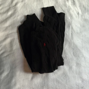 Svart kabelstickad tröja Polo Ralph Lauren - Svart kabelstickad tröja från Polo Ralph Lauren i storlek M. Klassisk rund hals och långärmad modell med ribbade muddar. Tröjan har en röd broderad logga på bröstet och är tillverkad i mjuk bomull. Perfekt för dig som gillar stilrena och tidlösa plagg. Kan passa folk med S. Har ett hål i ryggen som har blivit sytt men kan fortfarande synas, extremt enkelt att sy över mer så att hålet syns mindre. Pris kan diskuteras!
