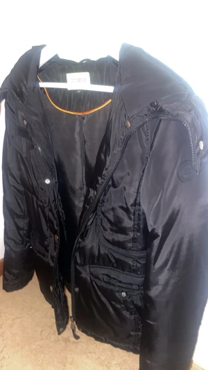 Roco Baroco - svart vinterjacka   - Svart vinterjacka från Roco Baroco med flera fickor, dragkedja och tryckknappar. Jackan har en hög krage och quiltade detaljer som ger en cool look. Materialet är glansigt syntet och jackan är långärmad, perfekt för kalla dagar under hösten och vintern!