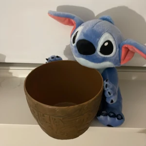 Stitch nalle som håller i en skål  - Bilderna visar en Stitch-mjukisdjur och en brun kruka med texten 'Aloha' och dekorativa mönster. Båda produkterna ser ut att vara i gott skick utan synligt slitage. Perfekt för Disney-fans eller som dekoration. Observera att detta inte är elektronik.