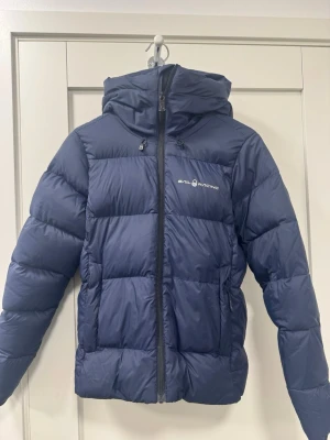 Marinblå Sail Racing dunjacka S - Säljer en marinblå dunjacka från Sail Racing med 700 fill power. Jackan har huva, två sidofickor med dragkedja och logga på bröstet samt på huvan. I bra skick. Inga hål eller andra skador.