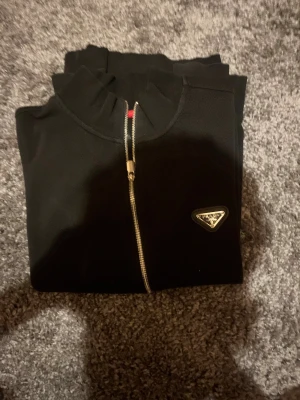 Svart ziptröja från Prada - Svart långärmad ziptröja från Prada med klassisk logga på bröstet. Tröjan har hel dragkedja i silver, ståkrage och två fickor framtill. Materialet är mjukt och känns som bomull, Använd i ungefär 1 år men väl omhändertagen