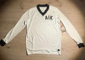AIK 1924 Edition Målvaktströja  - Snygg AIK fotbollströja från Nike i vit Dri-Fit polyester med svart V-ringad krage och muddar. Tröjan har broderad Nike-logga och AIK-tryck på bröstet, samt texten 'Allmänna Idrottsklubben' i nacken. Slim fit-modell som passar perfekt för match eller träning. Original Pris: 1299kr Skriv om ni har några funderingar kring tröjan
