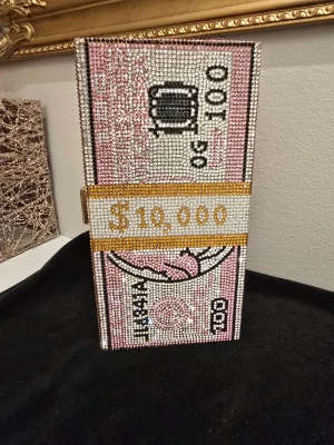 Glittrig kuvertväska dollar design - Kuvertväska i form av en bunt dollarsedlar, täckt med glittrande paljetter i rosa, vit, guld och svart. Väskan har rektangulär form och stängs med ett guldfärgat spänne på sidan. Perfekt statement piece för dig som gillar att sticka ut.