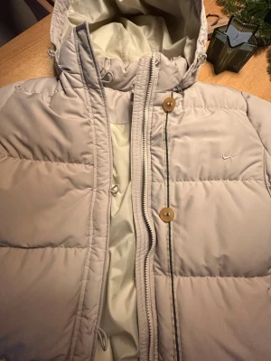 Ljusbeige lång dunjacka från Nike - Lång, ljusbeige dunjacka från Nike med huva, dragkedja och knappar framtill. Jackan är quiltad och har en clean look med diskret Nike-logga på bröstet. Perfekt för kalla dagar och riktigt snygg till vintern.