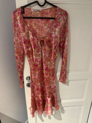 Blommig kortklänning från Zara - Klänning från Zara i rosa och orange toner. Klänningen har lång ärm, v-ringning med knyt framtill och volang nertill. Använd ett fåtal gånger. 