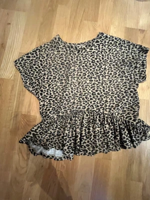 Leopardmönstrad topp från Kappahl - Säljer en snygg topp från Kappahl i storlek 146/152 med leopardmönster i beige och svart. Toppen har korta ärmar, rund hals och en volang nertill som ger en extra cool touch. Perfekt för dig som gillar djurmönster och vill sticka ut lite extra.