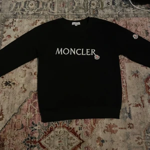 Svart sweatshirt från Moncler - Svart sweatshirt från Moncler med stor vit broderad logga på bröstet och patch på ärmen. Tröjan har rund hals, långa ärmar och ribbade muddar. Perfekt för dig som gillar stilrena och exklusiva märken.