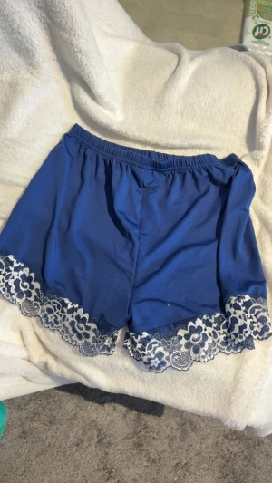 Blå shorts med spetskant - Snygga blå shorts med elastisk midja och dekorativ vit spetskant nertill. Perfekta för en chill sommarlook eller som mysplagg hemma. Mjukt material som känns skönt mot huden och ger en avslappnad vibe.