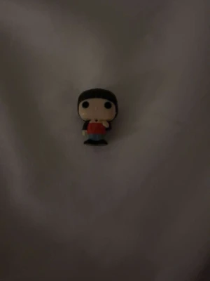 Röd väst på minifigur - Minifigur med en röd väst, svart hår och blå byxor. Västen har en enkel och klassisk form utan synliga detaljer som knappar eller dragkedja. Perfekt som dekoration eller samlarobjekt för dig som gillar små figurer med cool stil.