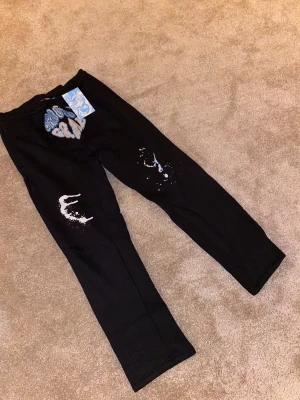 Gravity Grab Blackout Splash mjukisbyxor - Svarta mjukisbyxor från Gravity Grab med broderad logga i blått och grått framtill samt vita splash-detaljer på benen. Byxorna har snörning i midjan och är tillverkade i mjukt material för maximal komfort. Snygg streetwear vibe och unik design. Limted edition!!