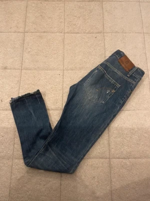 Blå dondup jeans - Säljer ett par blå jeans från dondup med klassisk femficksdesign, slitningar på knäet och råa benslut. Jeansen har en snygg tvätt och smal passform som sitter tajt längs benen. Perfekta för dig som gillar en avslappnad men trendig look.