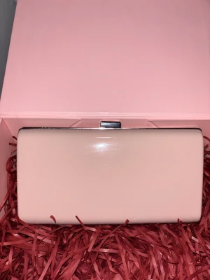 Ljusrosa kuvertväska från Anna Field - Stilren ljusrosa kuvertväska från Anna Field med silverfärgad ram och kedja. Väskan har en hård, slät yta och ett klassiskt metallspänne upptill. Perfekt att bära i handen eller över axeln med kedjan. Enkel och elegant design.