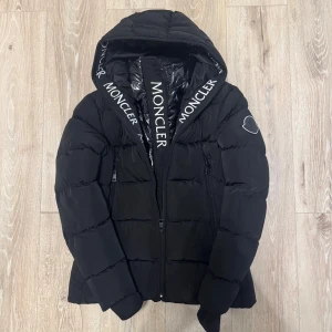 Svart Moncler pufferjacka med huva - Svart pufferjacka från Moncler med stor huva och logotyp på ärmen. Jackan har tvåvägsdragkedja, quiltad design och Moncler-tryck på insidan av huvan. Perfekt för kalla dagar med sin fluffiga och stilrena look.