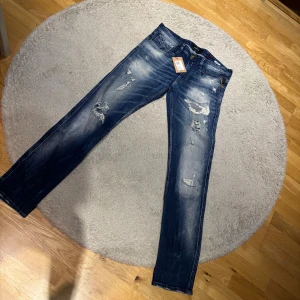 Replay Anbass blå slitna jeans - Snygga blå Replay Anbass jeans med slitningar och distressed detaljer på benen. Modellen är slim och har klassisk femficksdesign, med läderpatch bak i midjan. Jeansen har en mörk tvätt med ljusa partier och är tillverkade i stretchigt denim och Slim fit