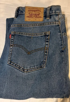 Levi's 501 blå jeans straight fit - Klassiska Levi's 501 jeans i ljusblå tvätt med raka ben och normal passform. Jeansen har fem fickor, knappgylf och den ikoniska läderpatchen bak i midjan. Tillverkade i slitstarkt bomullsjeansmaterial med orange sömmar och röd Levi's-tag på bakfickan.
