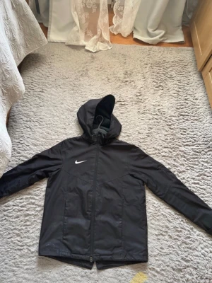 Svart vindjacka från Nike med huva - Svart vindjacka från Nike med klassisk logga på bröstet. Jackan har huva, hel dragkedja framtill och långärmade ärmar. Materialet är lätt och syntetiskt, perfekt för blåsiga dagar. Enkel design utan mönster, med praktiska fickor och sportig look.