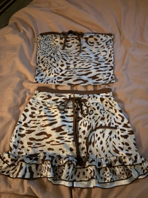 Leopardmönstrad bandeau tubtopp med kjol - Säljer en snygg bandeau tubtopp och kjol med leopardmönster i beige och brunt. Toppen har bruna kanter och en söt snörning framtill. Materialet är mjukt och stretchigt, perfekt för dig som gillar att sticka ut. Matcha med kjolen för en komplett look!