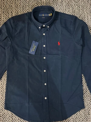 Mörkblå skjorta från Ralph Lauren - Klassisk mörkblå skjorta från Ralph Lauren med röd broderad logga på bröstet. Skjortan har button-down krage, vita knappar och är tillverkad i 100% bomull. Perfekt för dig som gillar stilrena och tidlösa plagg.