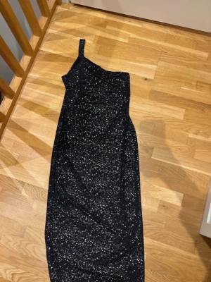 Svart glittrig långklänning oneshoulder - Svart långklänning med glittrigt mönster i silver. Klänningen har en snygg oneshoulder-design med bred axelrem och figurnära passform. Materialet är mjukt och stretchigt, perfekt för att sticka ut med en cool look. Mönstret ger en stjärnhimmel-vibe och gör klänningen extra unik. 