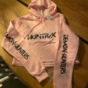 HUNTRIX K-pop Demon Hunters hoodie S - Säljer en ljusrosa crop huvtröja med kattöron i HUNTRX KPop Demon Hunters produktseries. Med tryck 'DEMON HUNTERS' på ärmarna och coolt Demon Hunters-motiv på ryggen.  För flicka/tjej. HELT NY med lapp, oanvänd pga köp av fel storlek. Vänligen kolla mått nedan noggrant innan du köper!  Storlek S. Mått: byst 49 cm. Axelbredd 43 cm. Armlängd 61 cm. Längd 43 cm.   