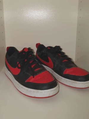 Nike Dunk Low svart/röd sneakers - Säljer ett par Nike Dunk Low sneakers i svart och rött med klassisk röd swoosh på sidan. Skorna har snörning, platt sula och rund tå. Ovandelen är i skinn och syntet, med vit mellansula och röd yttersula. Perfekt för dig som gillar streetstyle och sportig look.