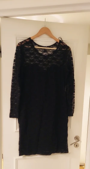 Svart spetsklänning från H&M - Svart kortklänning från H&M i storlek L, helklädd i blommig spets med långärmade ärmar. Klänningen har rund halsringning och transparenta spetsdetaljer upptill och på ärmarna. Perfekt för dig som gillar en stilren och elegant look.