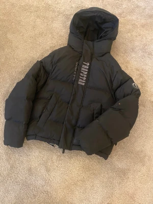 Svart pufferjacka från Trapstar - Säljer en svart pufferjacka från Trapstar äkta med stor broderad logga på ryggen och huvan. Jackan har huva, dragkedja och coola detaljer på ärmen. Perfekt för kalla dagar när du vill ha streetstyle-vibe. !äkta!