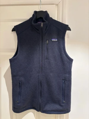 Mörkblå fleeceväst från Patagonia - Snygg mörkblå fleeceväst från Patagonia med hög krage och hel dragkedja framtill. Västen har tre praktiska fickor med dragkedja och ett diskret logomärke på bröstet. Perfekt för lager-på-lager och chill dagar.