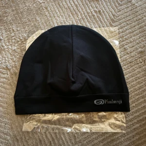 Svart träningsmössa från Kalenji - En stilren svart mössa från Kalenji, perfekt för träning och löpning. Mössan har en enkel, rundad form och är tillverkad i mjukt syntetmaterial som andas. Diskret Kalenji-logga i silver på kanten. Sitter skönt och passar dig som vill ha en clean look.