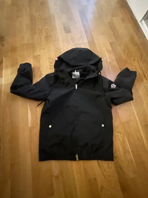 Svart vindjacka från Moncler - Svart vindjacka från Moncler med huva och dragkedja framtill. Jackan har Moncler-logga på ärmen och två fickor med silverfärgade knappar. Huva med röd, vit och blå randig detalj. Perfekt för dig som gillar stilren och sportig look.