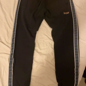 Svarta mjukisbyxor från Hugo Boss - Svarta mjukisbyxor från Hugo Boss med blå och vita sidoremsor där det står 'BOSS' längs hela benet. Byxorna har snörning i midjan och en slim passform. Perfekta för chill eller träning, riktigt snygga detaljer längs sidorna.