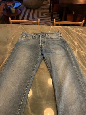 Levi's 501 ljusblå jeans W28 L32 - Klassiska Levi's 501 jeans i ljusblå tvätt med raka ben och normal passform. Jeansen har fem fickor, knappgylf och den ikoniska läderpatchen bak i midjan. Perfekt för dig som gillar tidlös denimstil och vill ha ett par jeans som funkar till allt.