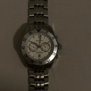 Silverfärgad kronografklocka från Gant - Snygg kronografklocka från Gant i silverfärgat rostfritt stål med länkarmband. Urtavlan är vit med två mindre subdials, datumvisning och tydliga markeringar. Klockan har vridbar ring och sportig design, perfekt för dig som gillar stilrena accessoarer.