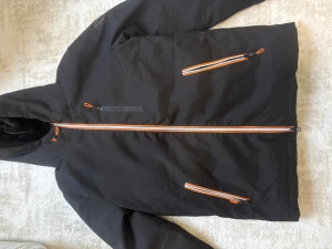 Svart dunjacka från GeoNorway med huva - Svart dunjacka från GeoNorway med huva och orange detaljer på dragkedjorna. Jackan har tre fickor med dragkedja och ett snyggt märke på ärmen. Insidan är quiltad och fodrad med mjuk svart päls i huvan. Perfekt för kalla dagar. Priset kan diskuteras.