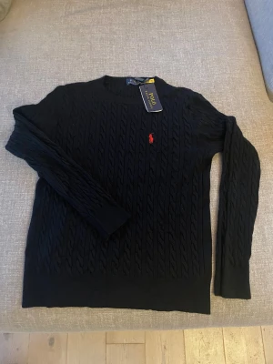 Svart kabelstickad tröja Polo Ralph Lauren - STORLEK: XS Svart kabelstickad tröja från Polo Ralph Lauren med rund hals och rött broderat logotyp på bröstet. Tröjan har långärmad design och ribbade muddar vid ärmslut och nederkant. Perfekt för dig som gillar klassisk stil med en modern touch.