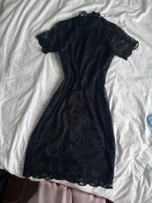 Svart spetsklänning med kort ärm - Säljer en svart, elegant spetsklänning med korta ärmar och hög hals. Klänningen har ett vackert blommigt spetsmönster och lätt figurnära passform. kanske passar bra till kontoret eller en begravning?hahahaha.