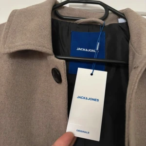 Beige rock från Jack & Jones Originals - Snygg beige rock från Jack & Jones Originals med klassisk krage och svarta knappar framtill. Jackan har en rak och loose passform, långa ärmar och är tillverkad i ett ullblandat material som ger en stilren look. Perfekt för dig som gillar minimalistisk och clean stil.