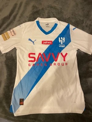 Vit och blå Al Hilal Puma fotbollströja - Säljer en officiell Al Hilal matchtröja från Puma i storlek M. Tröjan är vit med en diagonal blå rand och har klubbens logga på bröstet samt röda och blå tryck. Materialet är lätt polyester och har autentisk licensmärkning. Perfekt för dig som älskar fotboll och vill ha något unikt.