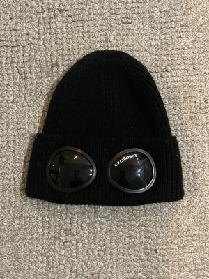 Svart C.P. Company Mössa - Svart ribbstickad mössa från C.P. Company med ikoniska goggles framtill. Snygg och unik design som sticker ut, perfekt för dig som vill ha streetwear-vibe. Mjuk och varm, tillverkad i stickat material. Nypris 1600kr.