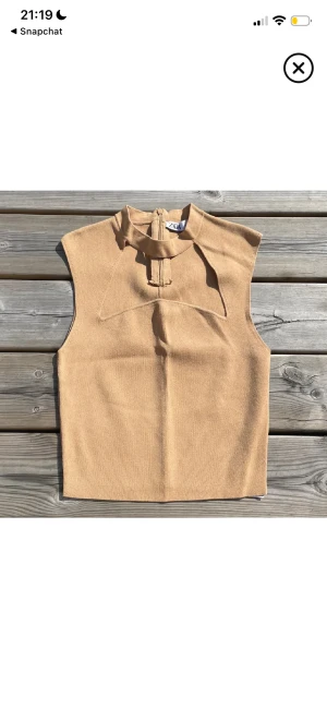Beige ärmlös topp från ZARA - Säljer en beige ärmlös topp från ZARA med hög hals och snygg detalj framtill. Toppen har en stilren, croppad passform och är tillverkad i ett mjukt syntetmaterial. Perfekt för dig som gillar minimalistisk och trendig stil.