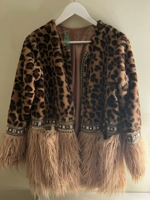 Leopardmönstrad pälsjacka med fransar - Unik pälsjacka med leopardmönster i bruna och beige toner, dekorerad med breda band med pärldetaljer och långa fluffiga fransar längst ner. Insidan är ljusbeige och jackan har en loose passform. Perfekt statement-plagg för dig som vill sticka ut. Finns inte någon storlek, men skulle beskrivas som en M