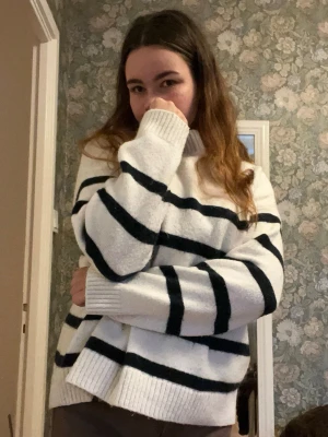 Vit och svart randig polotröja - Säljer en mysig vit polotröja med svarta horisontella ränder. Tröjan har en ribbad hög krage och långa ärmar med breda muddar. Materialet är stickat och ger en chill oversized vibe. Perfekt för dig som gillar klassisk stil med en twist.