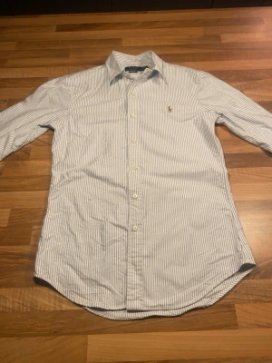Randig skjorta från Polo Ralph Lauren - Klassisk slim fit skjorta från Polo Ralph Lauren i vitt och blått med tunna vertikala ränder. Skjortan har lång ärm, knappar framtill och broderad logga på bröstet. Tillverkad i mjuk bomull och passar perfekt till en clean och stilren look.