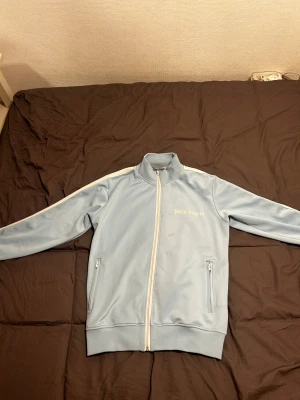 Ljusblå Palm Angels zip-tröja - Säljer en ljusblå zip-tröja från Palm Angels med vit dragkedja och vita ränder längs ärmarna. Tröjan har ståkrage, två fickor med dragkedja och Palm Angels-logga i guld på bröstet. Materialet är mjukt och känns som polyester, perfekt för en chill streetwear-look. Hör gärna av er om ni har frågor 