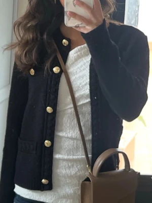 Svart kofta med guldknappar H&M  - Stilren svart kofta från H&M i stickad kvalitet. Koftan har rund halsringning, långa ärmar och snygga guldknappar framtill som ger en lyxig touch. Perfekt att slänga över en topp för en chic look. Passar dig som gillar klassiska detaljer.