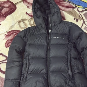 Svart dunjacka Sail Racing S - Svart dunjacka från Sail Racing i storlek S. Jackan har en stor huva, tydlig logga på bröstet och huvan, samt dragkedja framtill. Puffad design med breda kanaler och två sidofickor. Perfekt för kalla dagar och har en clean, sportig look.